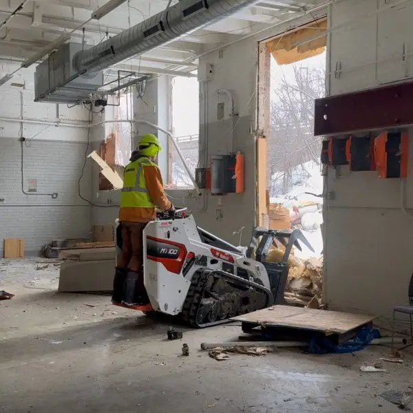 Interior demolition in progress, worker operating skid steer in prepared space for renovation.