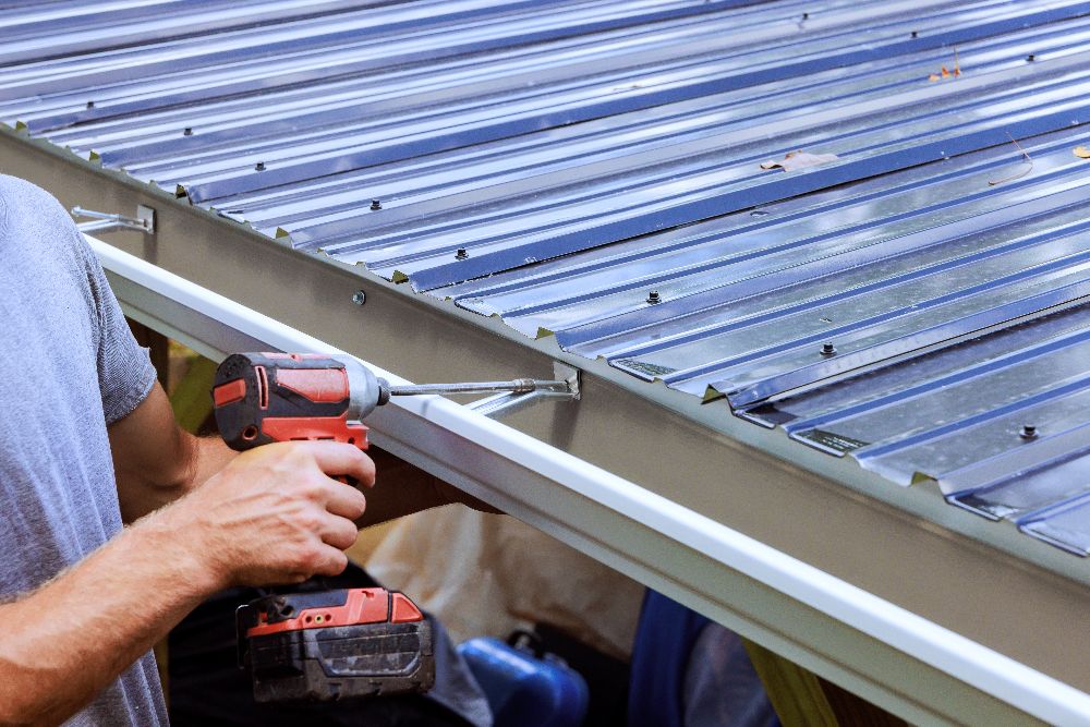 Installer using a power drill to secure a gutter along a metal roof edge